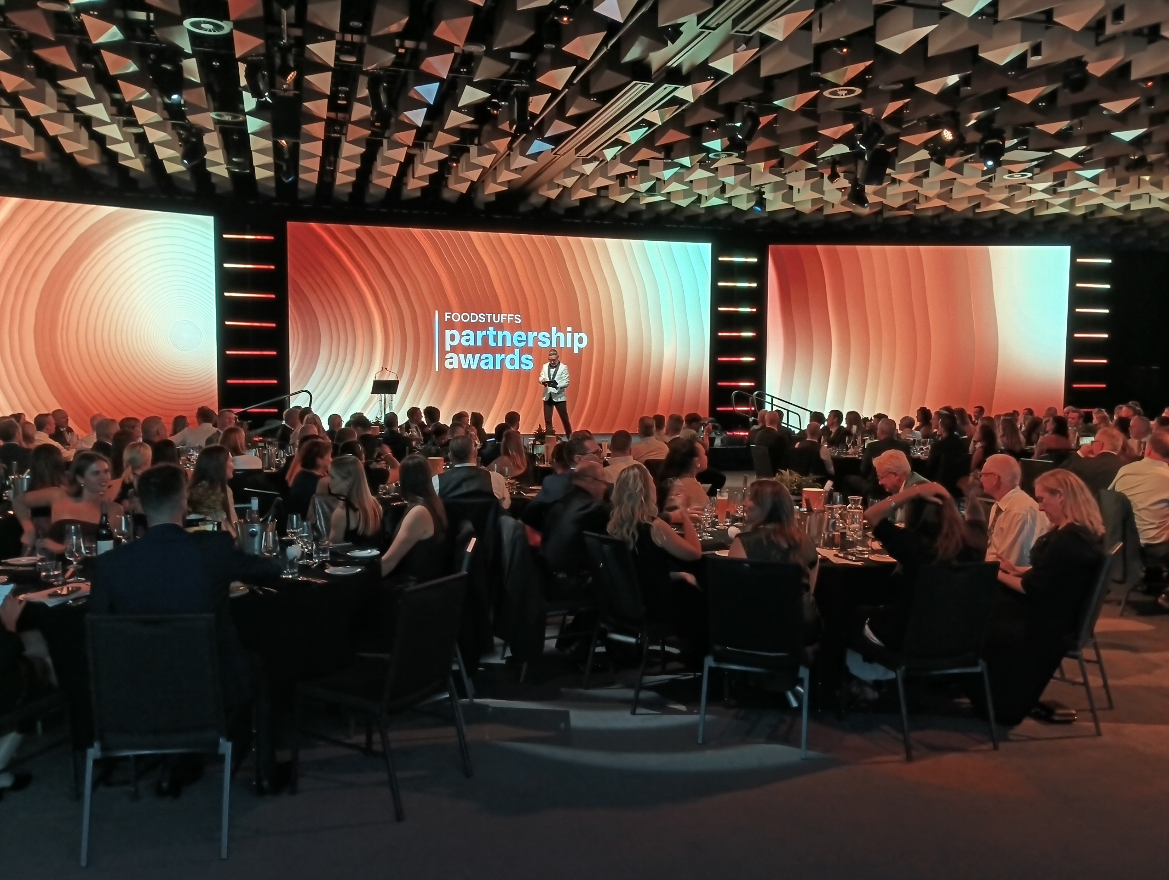 Packed awards dinner with warm amber stage screens and a presenter on stage in front of a seated audience.