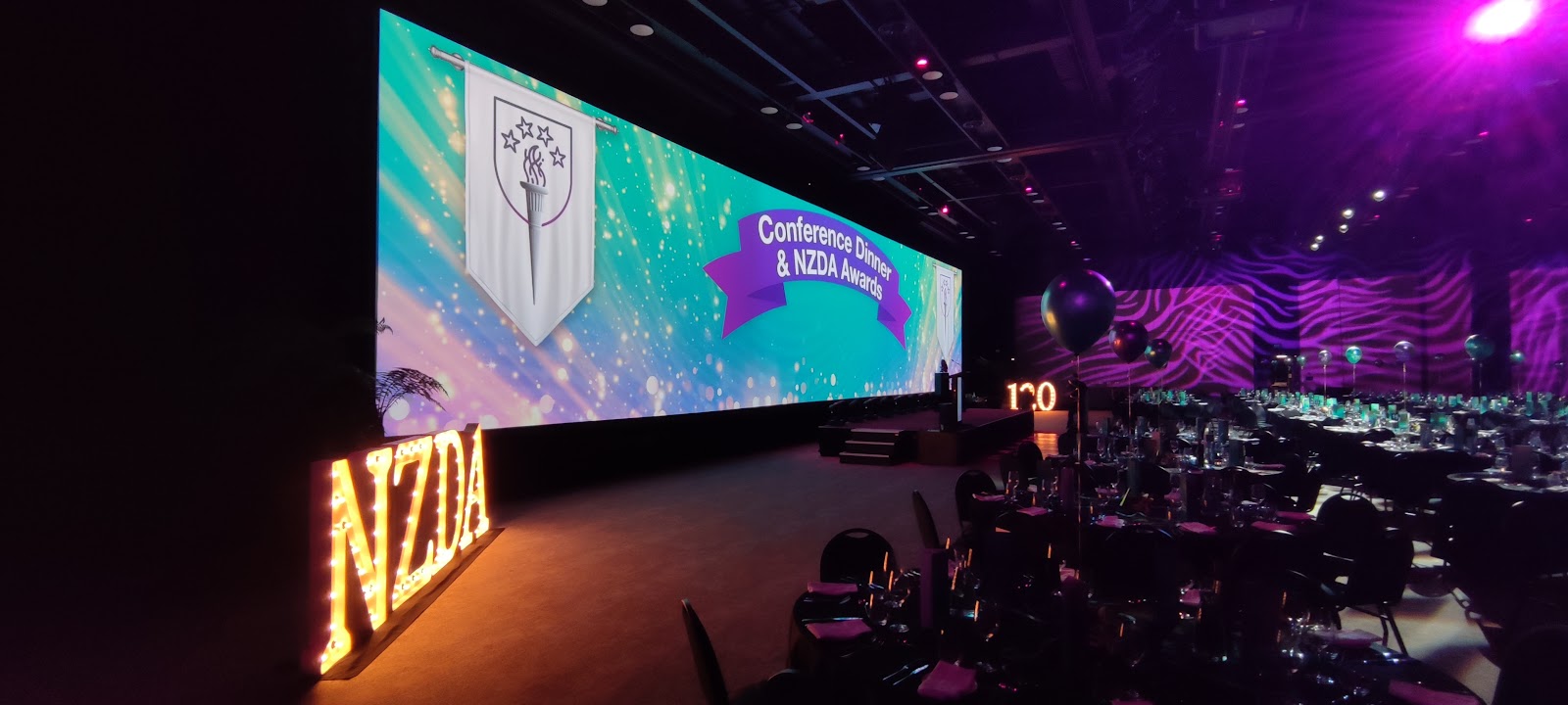 Awards dinner setup with a panoramic LED stage backdrop, lit NZDA letters, and banquet seating.